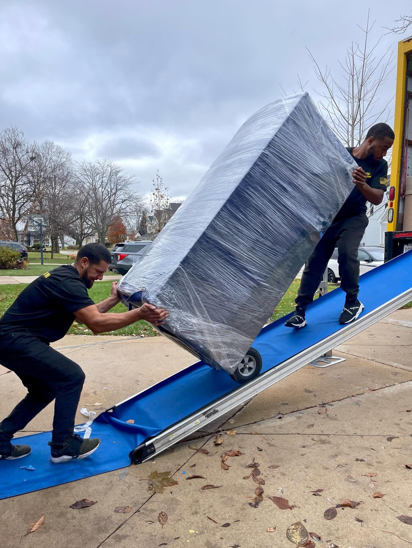 Yellow Truck Moving movers carefully loading a wrapped mattress up a truck ramp